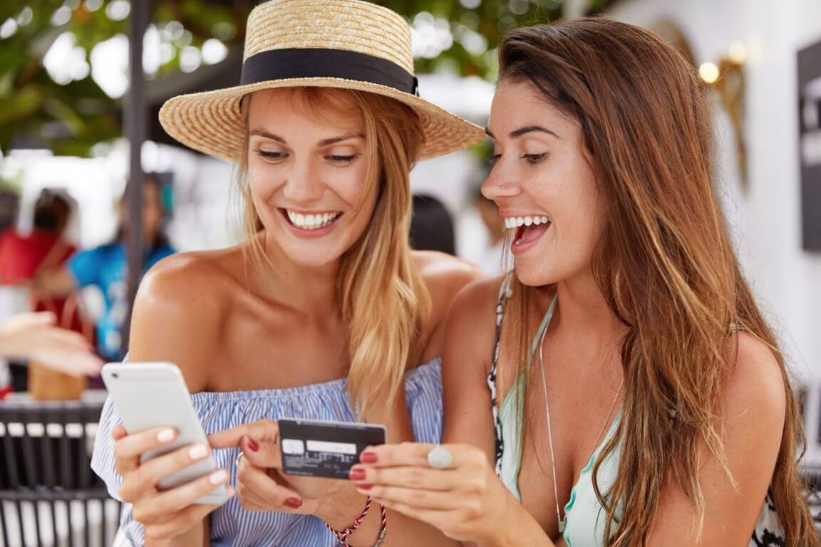 Cheerful best friends women meet together at cafeteria, happy to make online shopping with smart phone and plastic card, choose new clothes on internet website, happy to see something appropriate